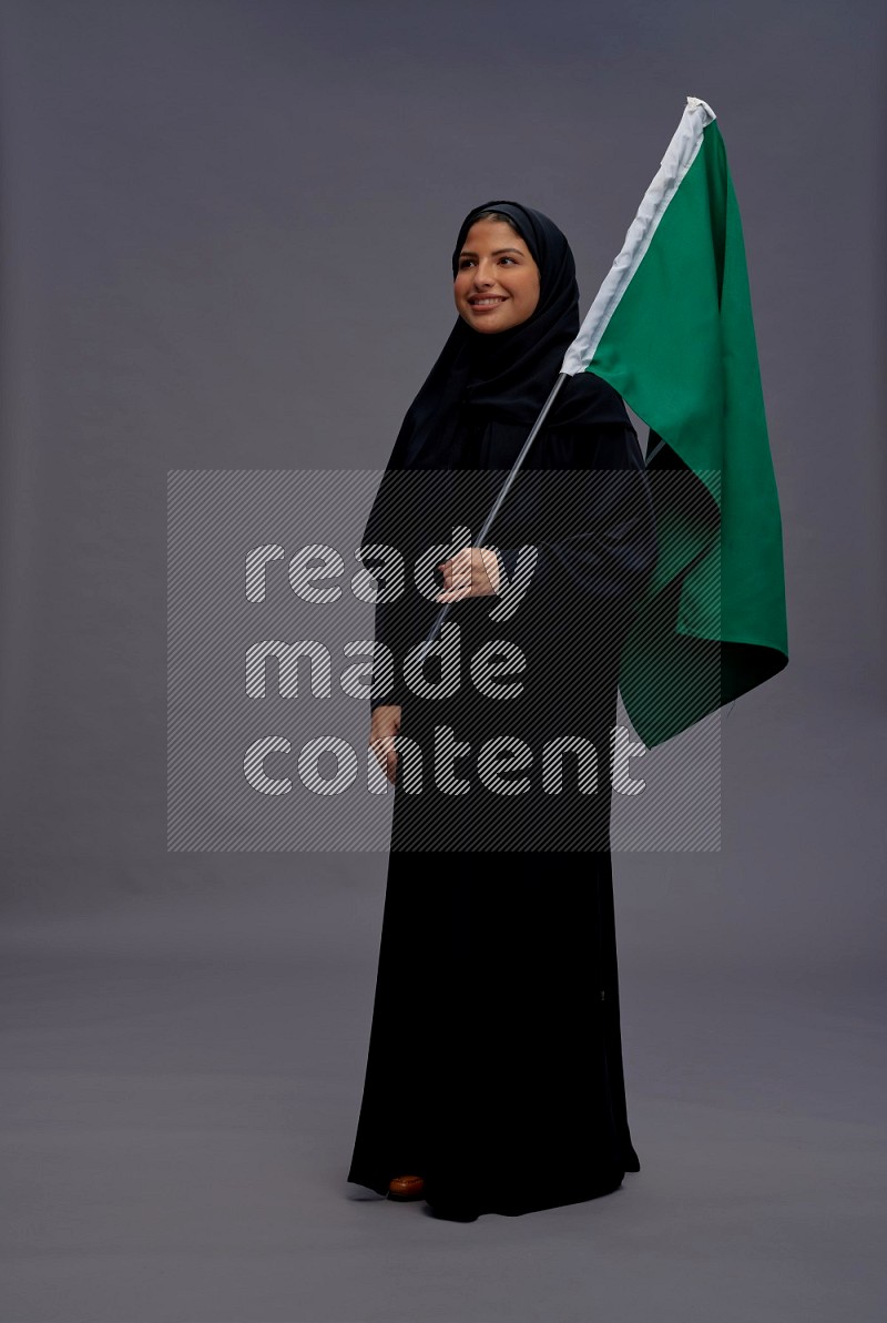 Saudi woman wearing Abaya standing holding Saudi flag on gray background