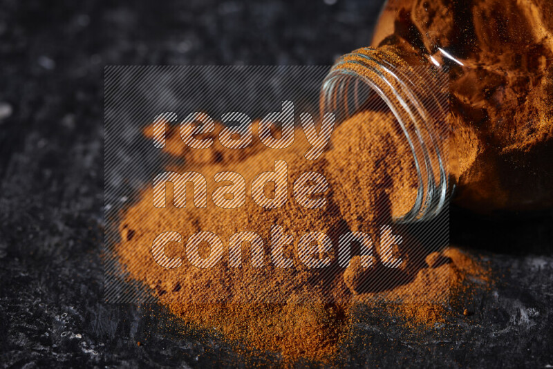 A glass jar full of ground paprika powder flipped with some spilling powder on black background