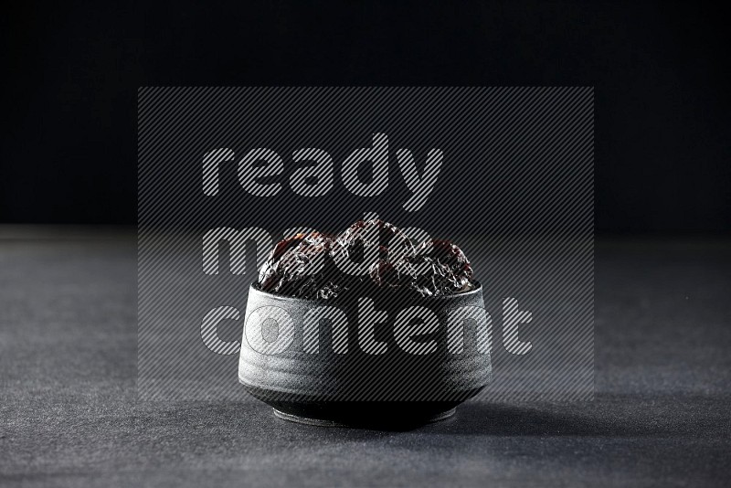 A black pottery bowl full of dried plums on a black background in different angles