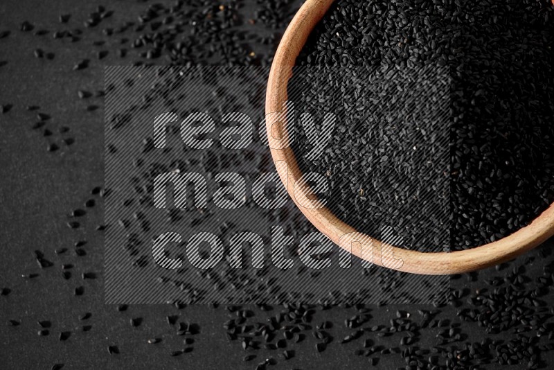 A wooden bowl full of black seeds surrounded by seeds on a black flooring