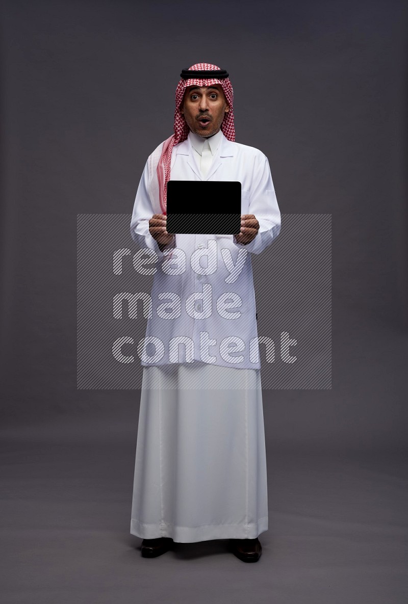 Saudi man wearing thob with lab coat and shomag with pocket employee badge standing showing tablet to camera on gray background