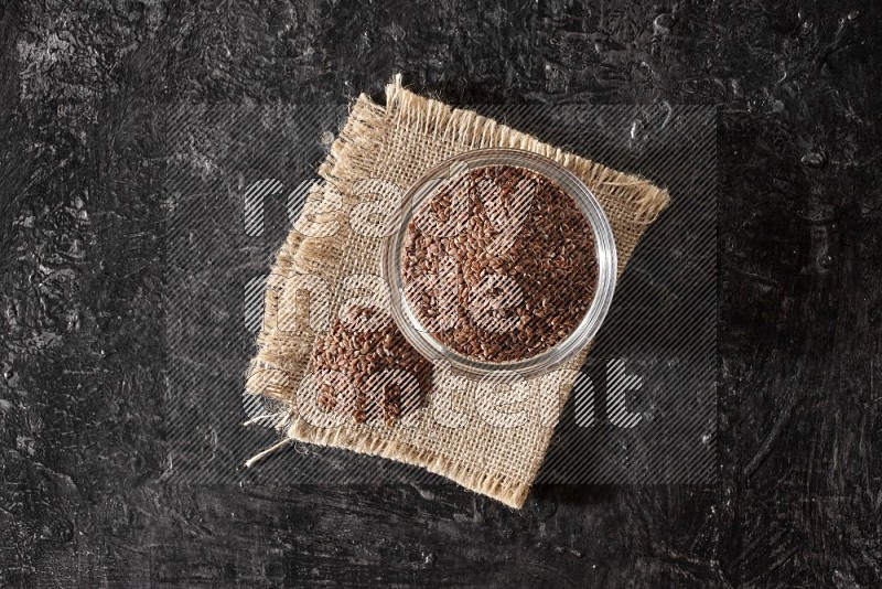 A glass bowl full of flax and seeds on burlap fabric on a textured black flooring in different angles