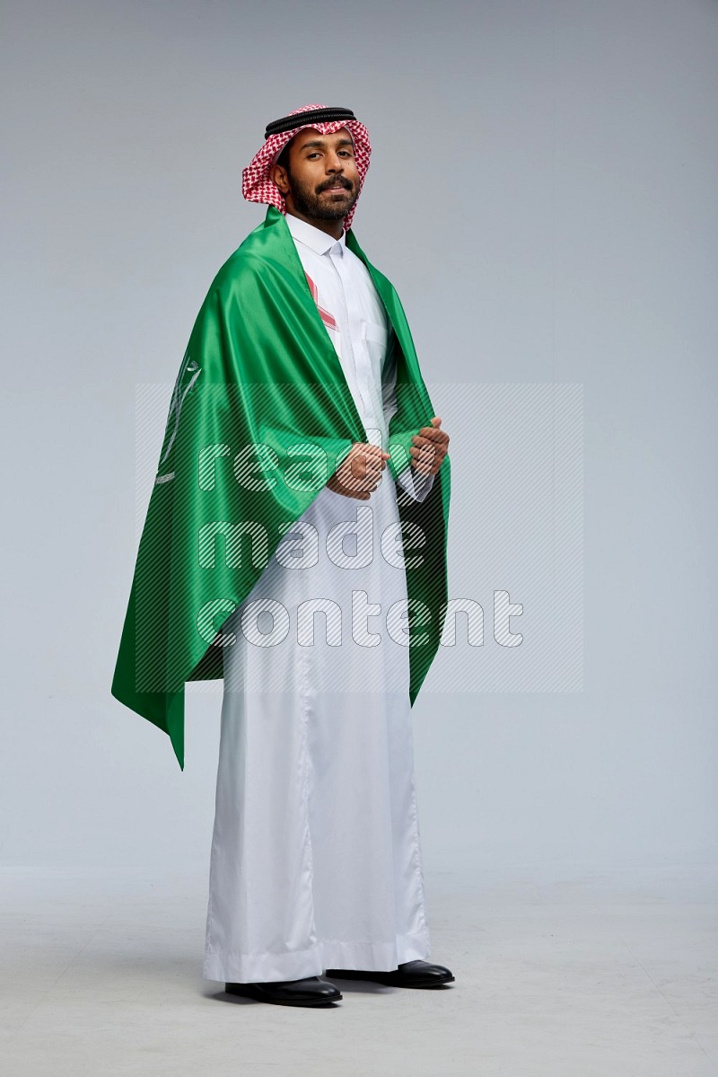 Saudi man Wearing Thob and shomag standing holding Saudi flag on Gray background