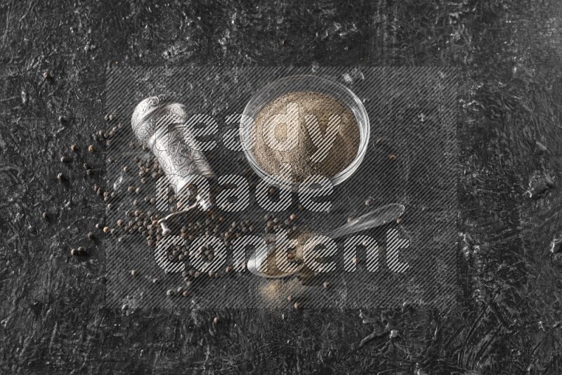 A glass bowl full of black pepper powder with black pepper beads, a turkish metal pepper grinder and a metal spoon on textured black flooring