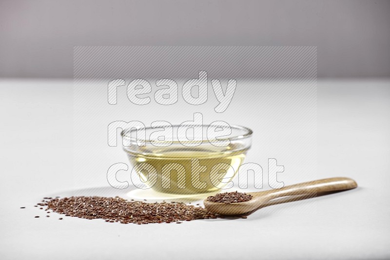 A glass bowl full of flaxseeds oil and wooden spoon full of flaxseeds with spread seeds on a white flooring