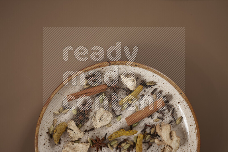 An assorted whole spices on a flat beige plate including fresh and dried ginger, cinnamon sticks, cardamom, turmeric fingers, cloves, star anise on a beige background