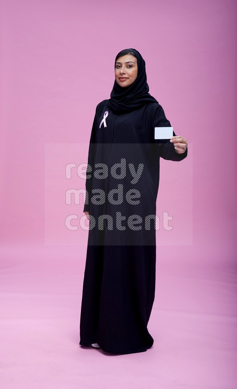 Saudi woman wearing pink ribbon on Abaya standing holding ATM card on pink background