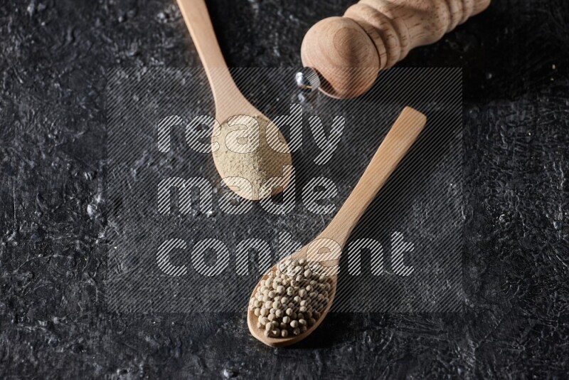 2 wooden spoons one full of white pepper powder and the other with pepper beads and a wooden pepper mill on textured black flooring