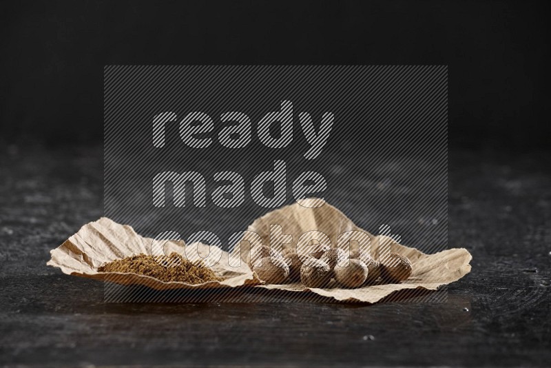 2 crumpled piece of paper full of nutmeg seeds and powder on a textured black flooring in different angles