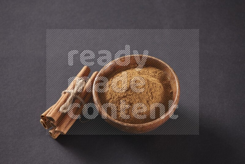 Cinnamon sticks stacked and bounded beside a wooden bowl full of cinnamon powder and a wooden spoon full of powder on black background