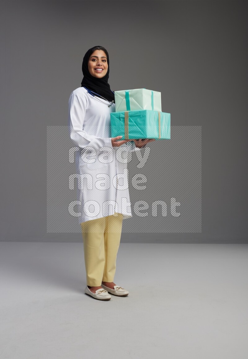 Saudi woman wearing lab coat with stethoscope standing holding gift box on Gray background