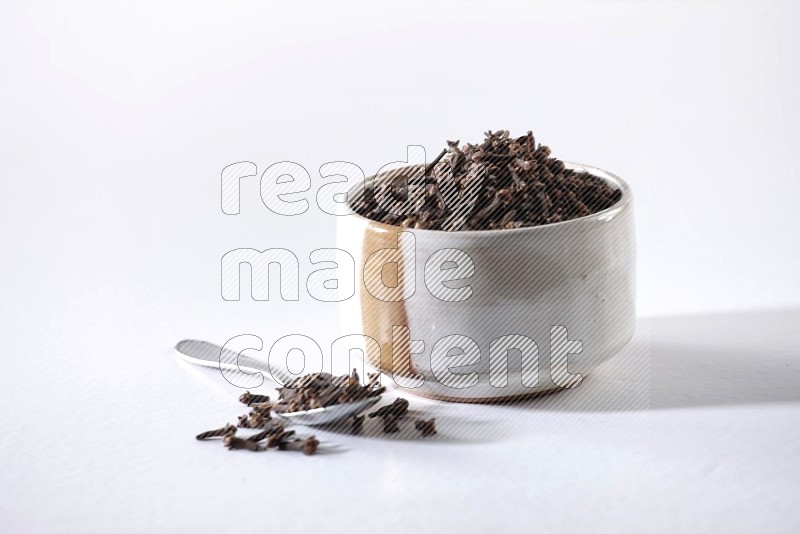 A beige ceramic bowl full of cloves and a metal spoon next to it on a white flooring