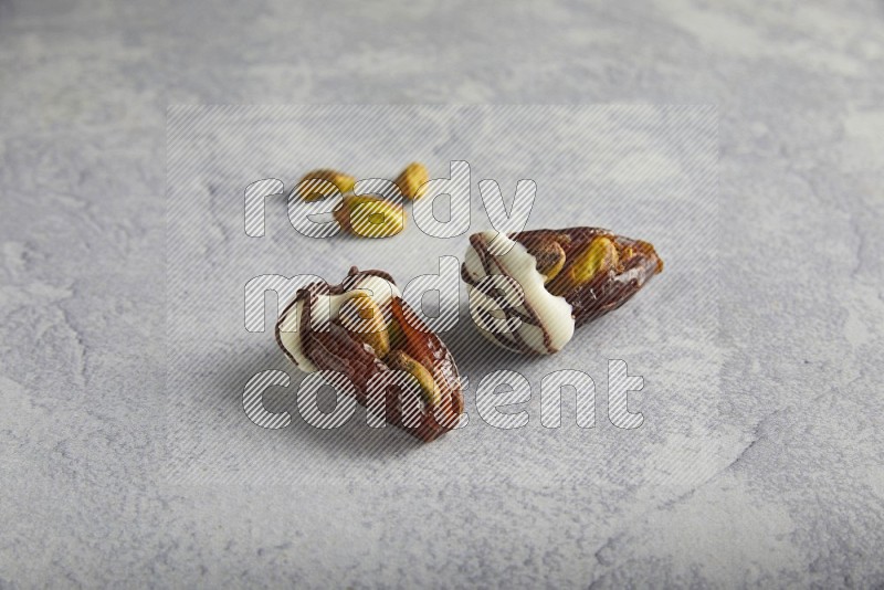 two pistachio stuffed dates covered with white chocolate with dark chocolate drizzle with unroasted pistachios on a light grey background