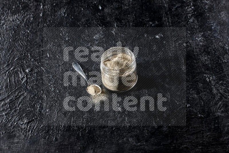 A glass jar and a metal spoon full of white pepper powder on textured black flooring