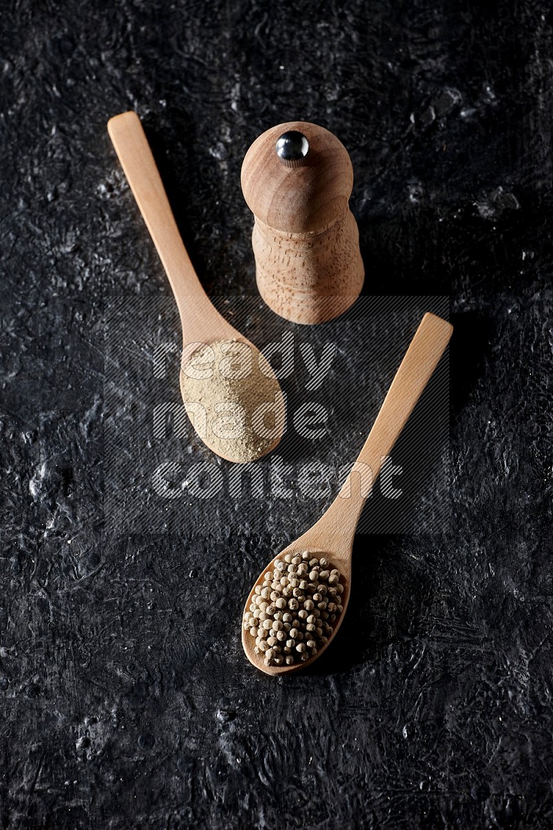 2 wooden spoons one full of white pepper powder and the other with pepper beads and a wooden pepper mill on textured black flooring