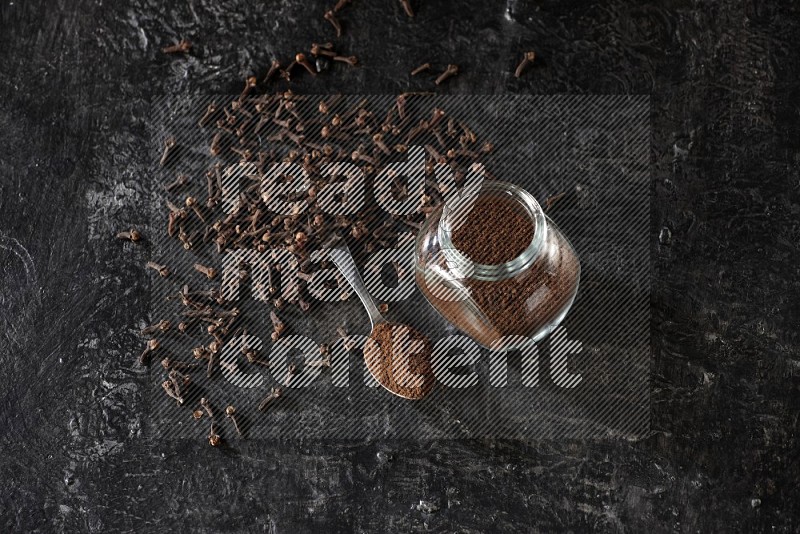 A glass spice jar and a metal spoon full of cloves powder with cloves spread on textured black flooring