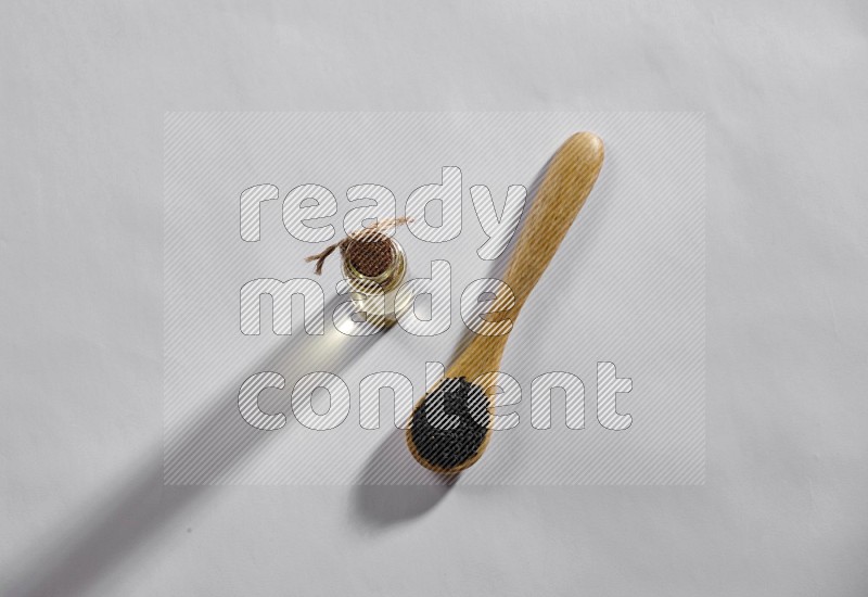 A wooden spoon full of black seeds with a glass bottle of black seeds oil on a white flooring