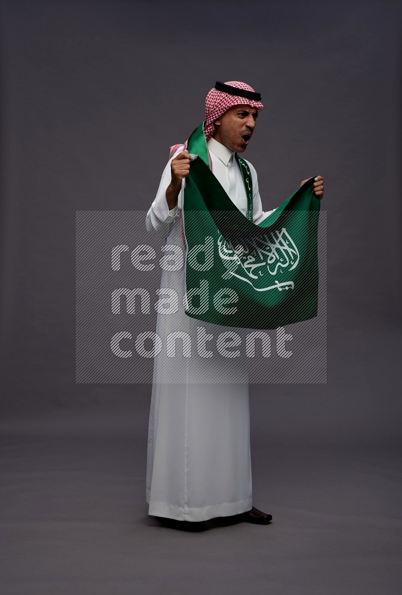 Saudi man wearing thob and shomag standing holding Saudi flag on gray background