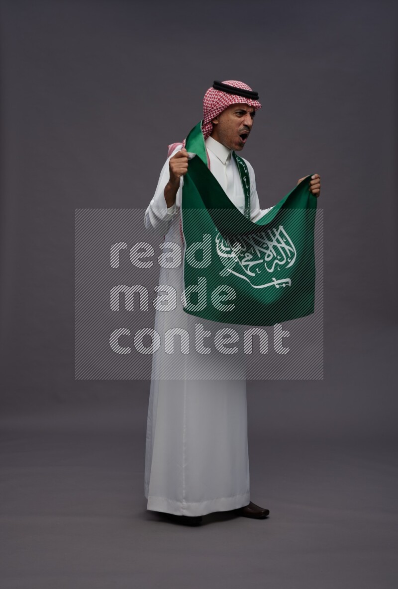Saudi man wearing thob and shomag standing holding Saudi flag on gray background