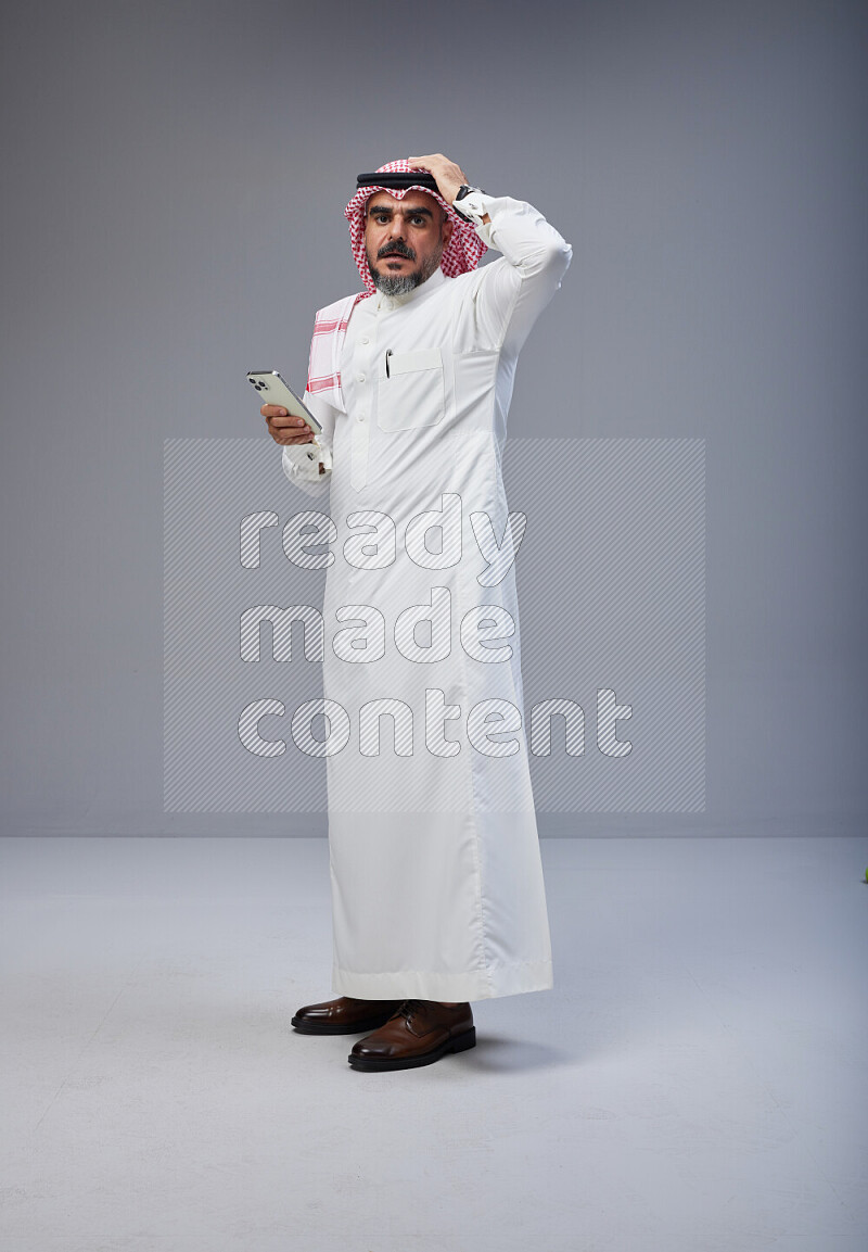 Saudi man Wearing Thob and red Shomag standing texting on phone on Gray background