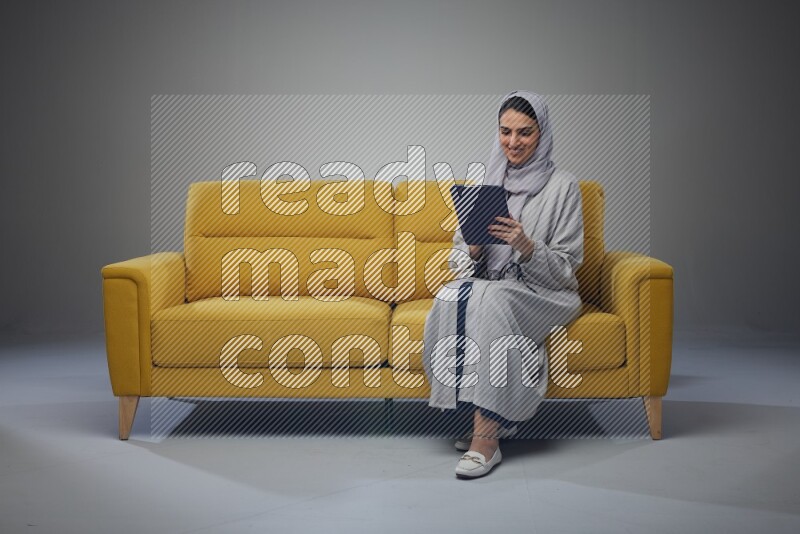 A Saudi woman wearing a light gray Abaya and head scarf sitting on a yellow sofa and holding her tablet while pointing to it eye level on a grey background