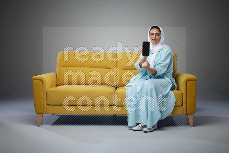 A Saudi woman wearing a light blue Abaya and white head scarf sitting on a yellow sofa and showing her phone's screen vertically and horizontally eye level on a grey background