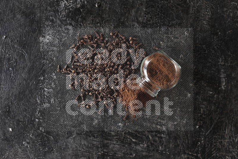 A flipped glass spice jar full of cloves powder and powder came out of it with cloves spread on textured black flooring