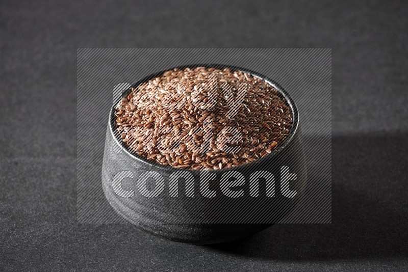A black pottery bowl full of flaxseeds on a black flooring