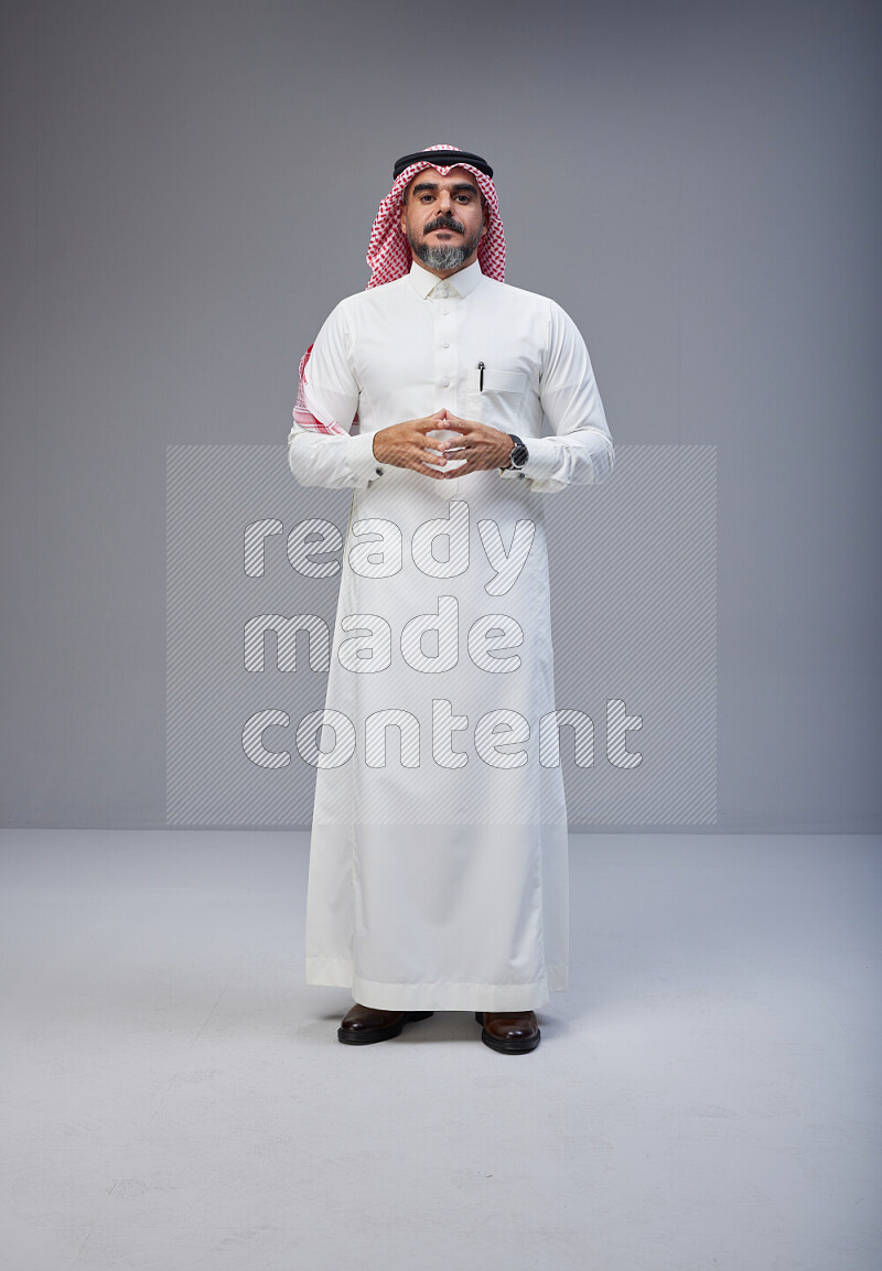 Saudi man Wearing Thob and red white Shomag standing interacting with the camera on Gray background