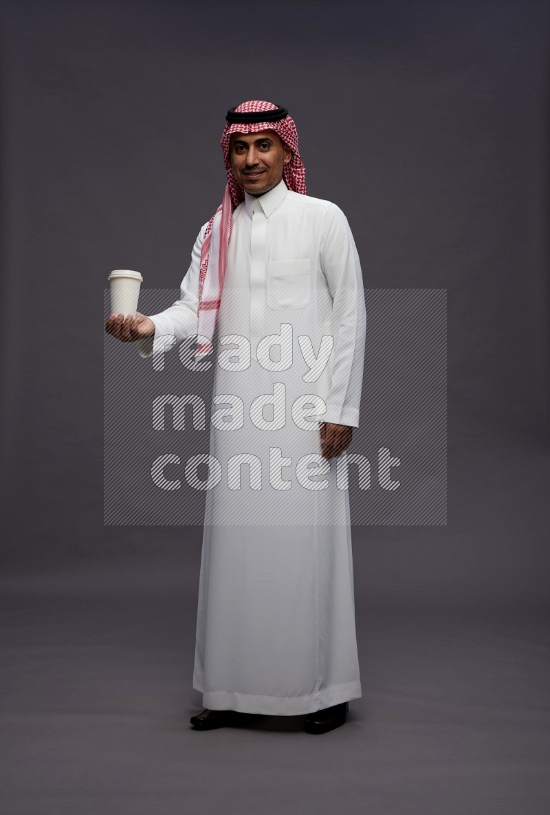 Saudi man wearing thob and shomag standing holding paper cup on gray background