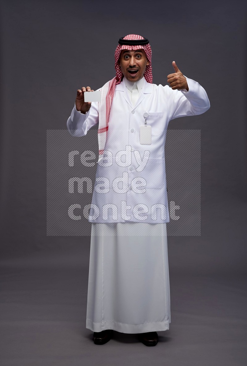 Saudi man wearing thob with lab coat and shomag with pocket employee badge standing holding ATM card on gray background