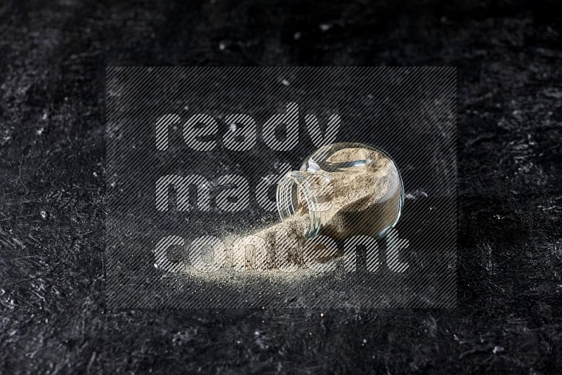 A flipped herbal glass jar full of white pepper powder with spilled powder on textured black flooring