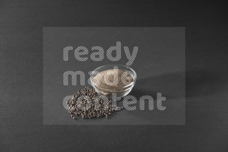 فلفل أسود مطحون في وعاء زجاجي وحبوب بجانبها علي خلفية سوداء