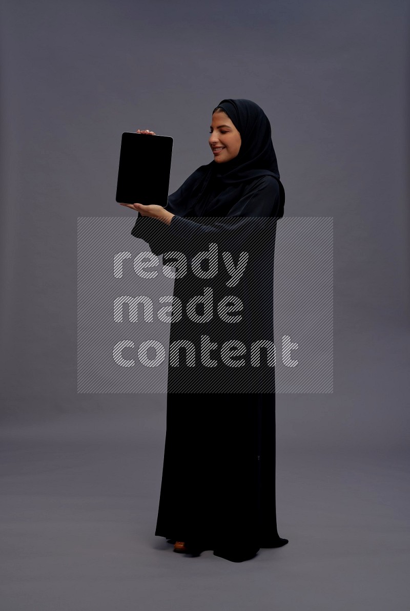 Saudi woman wearing Abaya standing showing tablet to camera on gray background