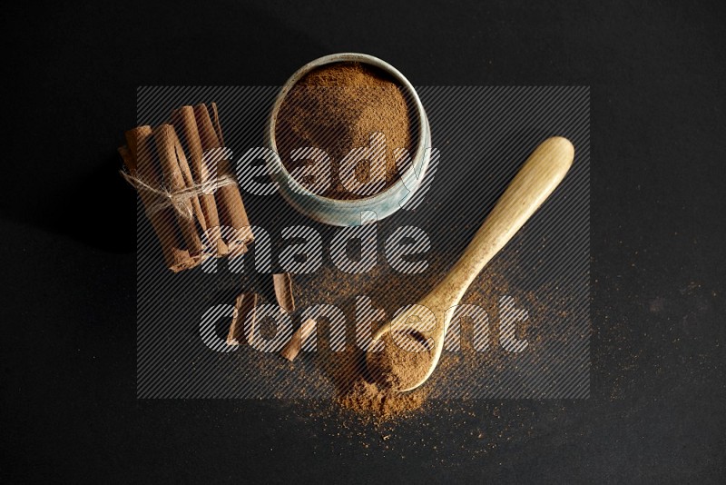 Ceramic bowl full of cinnamon powder and a wooden spoon full of powder with cinnamon sticks stacked and bounded on black background