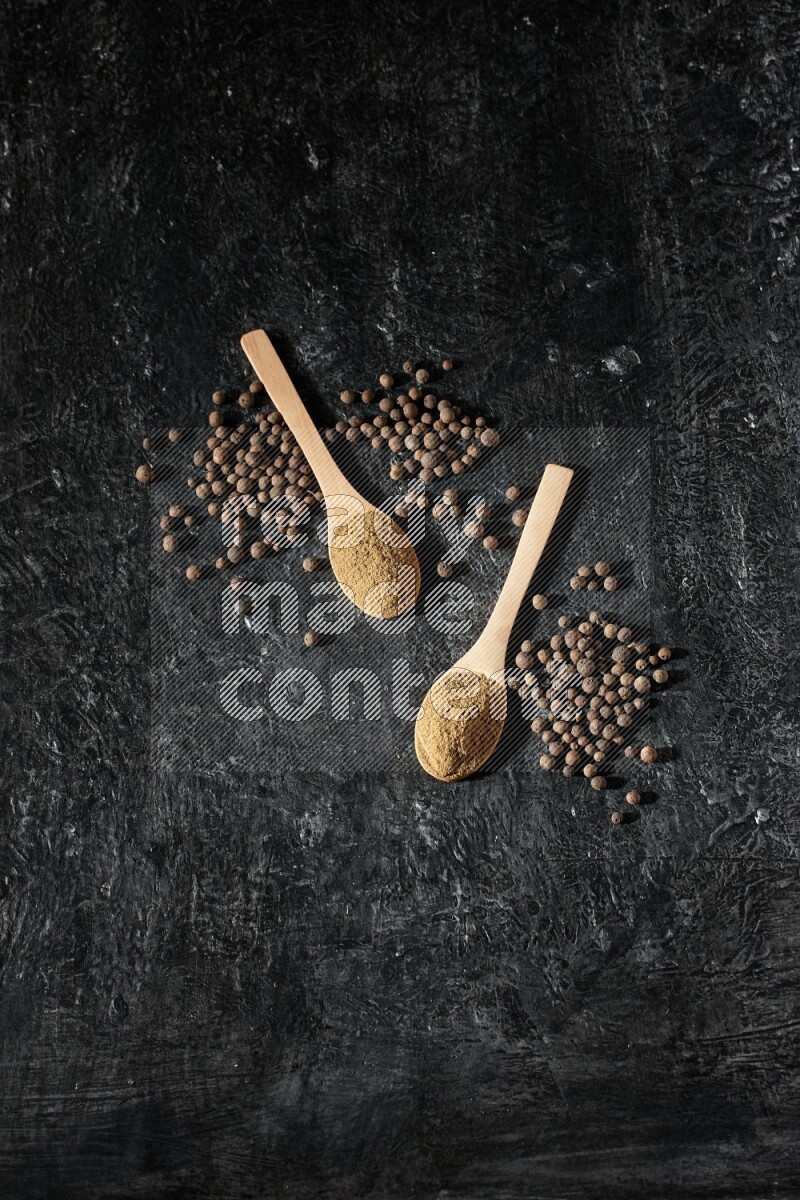 Wooden spoons full of all spice powder and allspice whole balls beside it on a textured black flooring