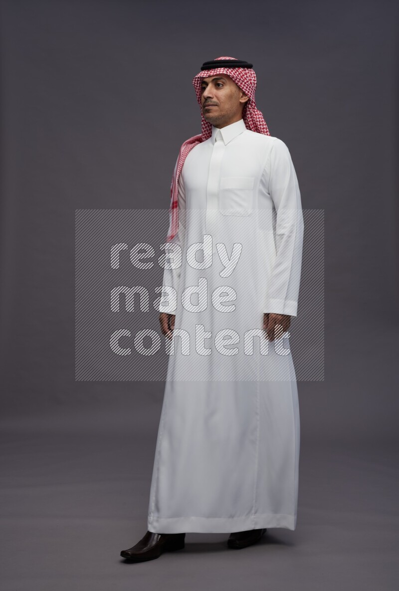 Saudi man wearing thob and shomag standing interacting with the camera on gray background