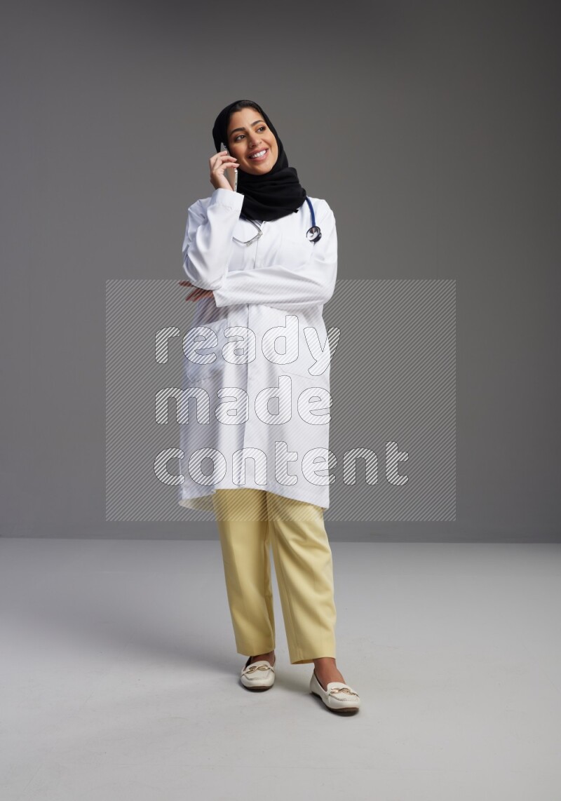 Saudi woman wearing lab coat with stethoscope standing talking on phone on Gray background