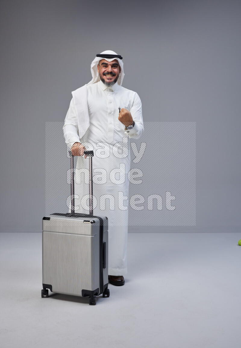 Saudi man wearing Thob and white Shomag standing holding Travel bag on Gray background