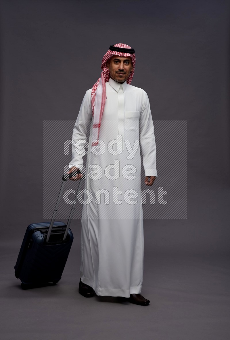 Saudi man wearing thob and shomag standing holding bag on gray background