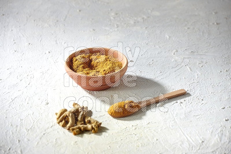A wooden bowl and wooden spoon full of turmeric powder with dried turmeric fingers on textured white flooring