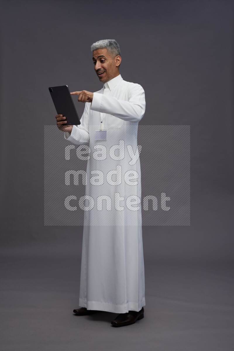 Saudi man wearing thob with neck strap employee badge standing working on tablet on gray background