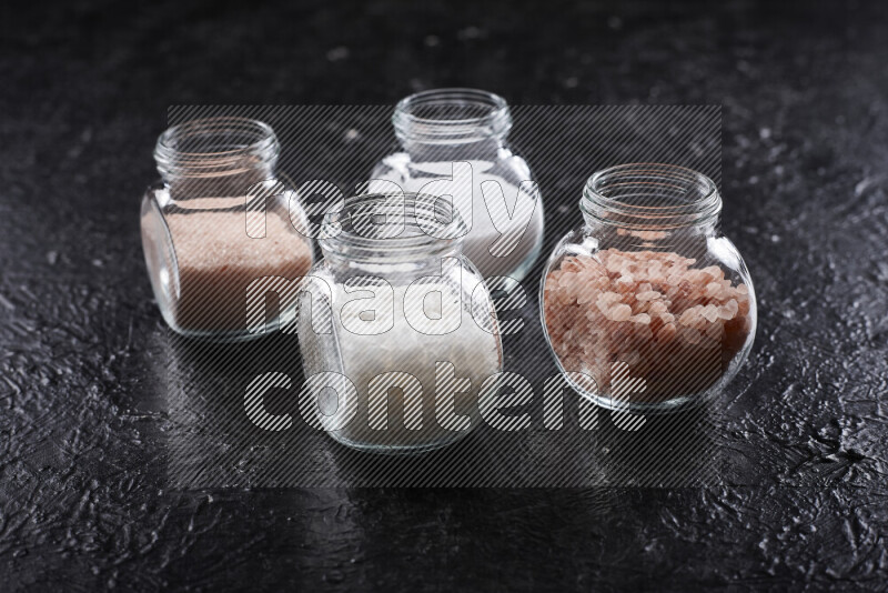 4 glass jars filled with table salt, coarse sea salt, fine himalayan salt and coarse himalayan salt on black background