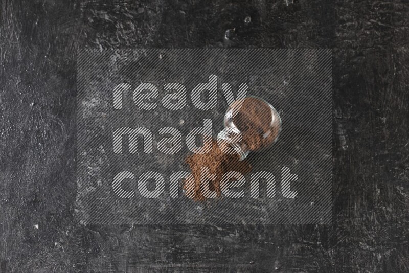 A flipped glass spice jar full of cloves powder and powder came out of it on textured black flooring