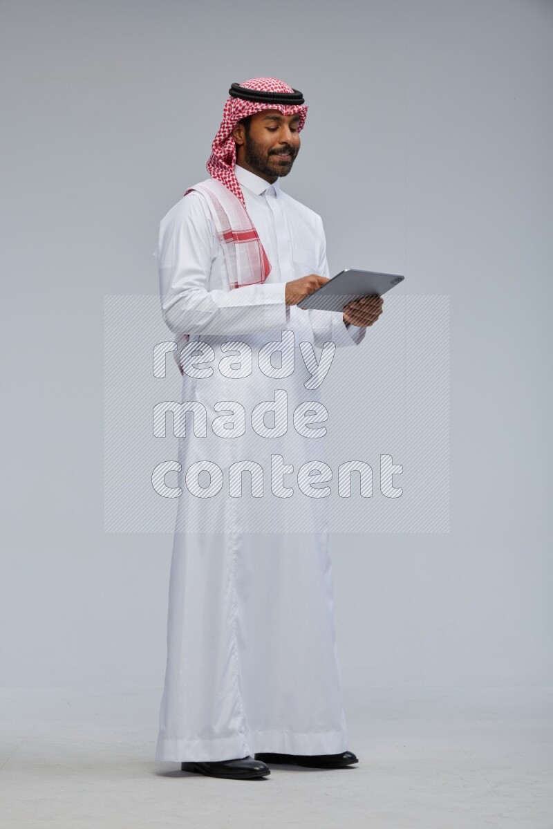 Saudi man Wearing Thob and shomag standing working on tablet on Gray background