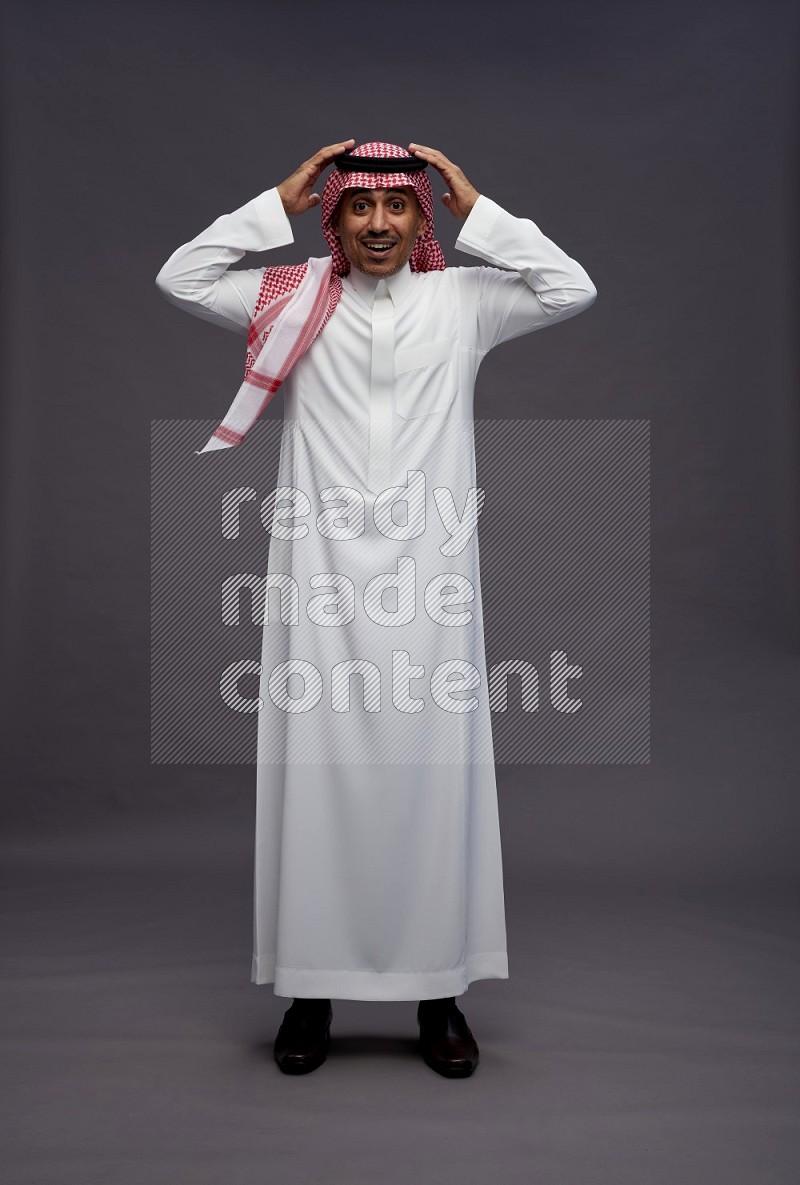 Saudi man wearing thob and shomag standing hands behind head on gray background