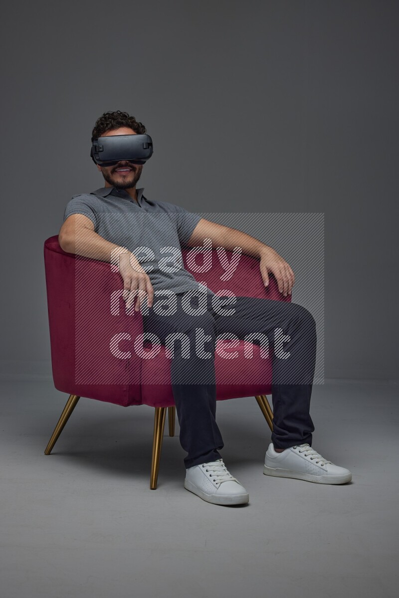 A man wearing casual and wearing VR while sitting on a burgundy chair eye level on a gray background