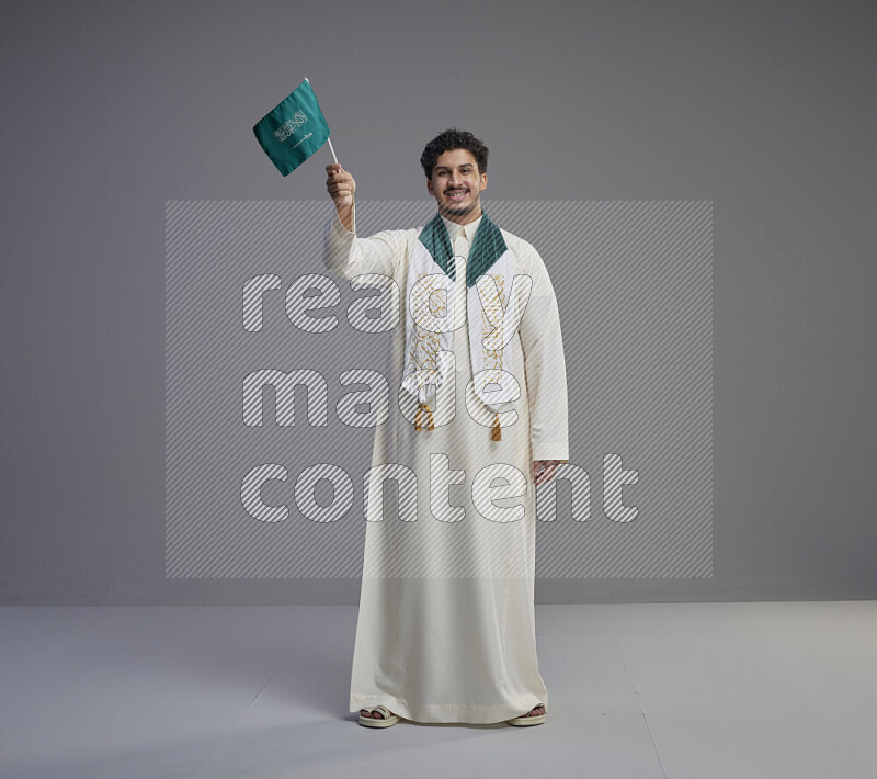 A Saudi man standing wearing thob and saudi flag scarf and holding small saudi flag on gray background