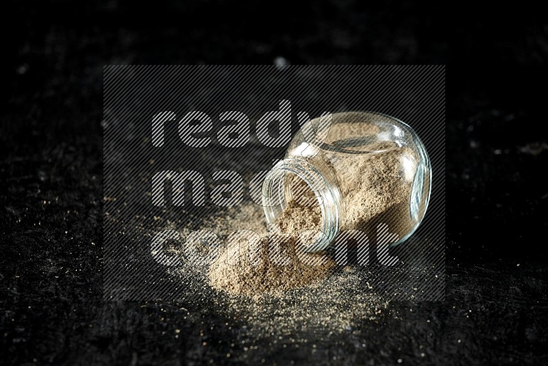 A flipped glass spice jar full of cardamom powder on textured black flooring