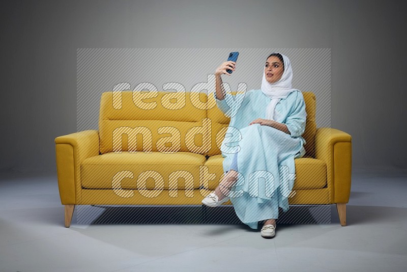 A Saudi woman wearing a light blue Abaya and white head scarf sitting on a yellow sofa and taking selfies with her phone eye level on a grey background